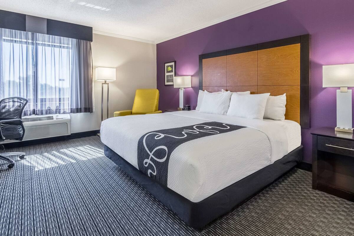 Ein Hotelzimmer mit einem großen Bett, einem Sessel, einem Schreibtisch und einem Fenster mit Vorhängen.