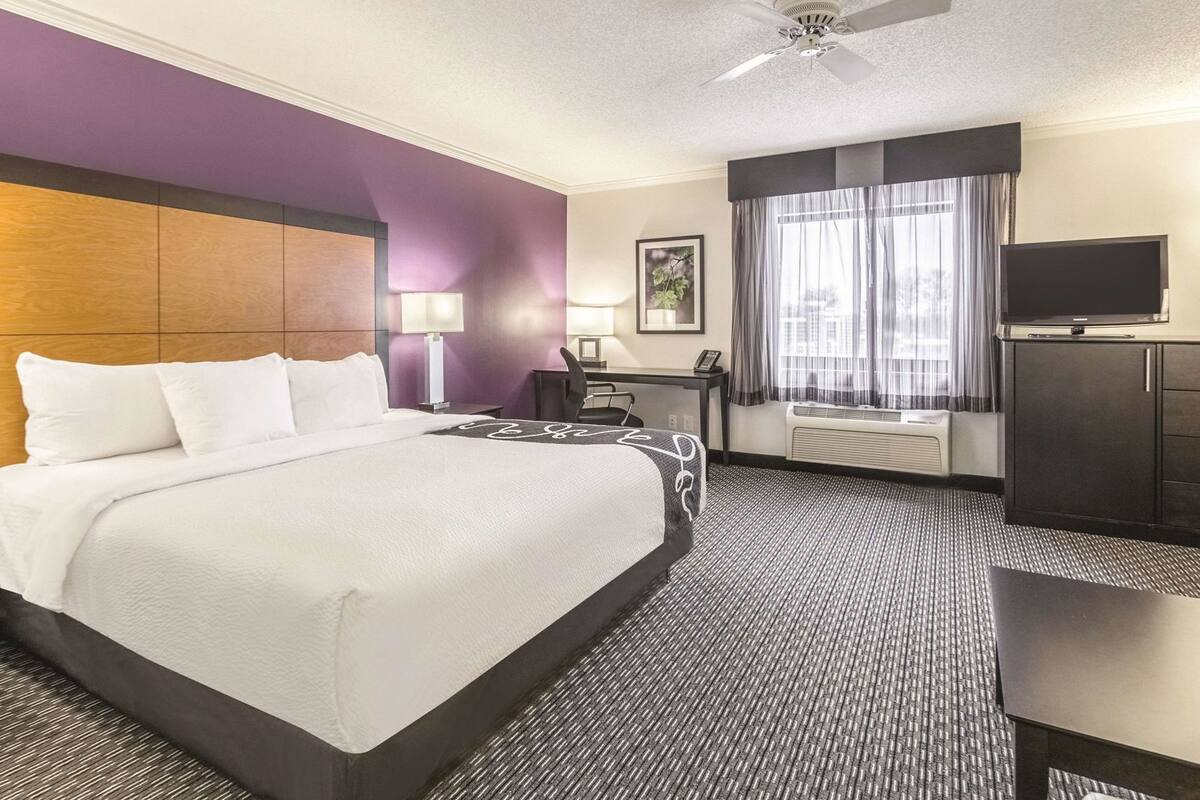Ein Hotelzimmer mit Bett, Schreibtisch, Sessel, Fernseher und Deckenventilator.