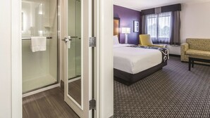 Habitación de hotel con mampara de vidrio, cama con colcha blanco y negro, sillón amarillo y ventilador de techo.