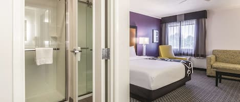A hotel room with a glass shower door, a bed with a black and white bedspread, a yellow armchair, and a ceiling fan.