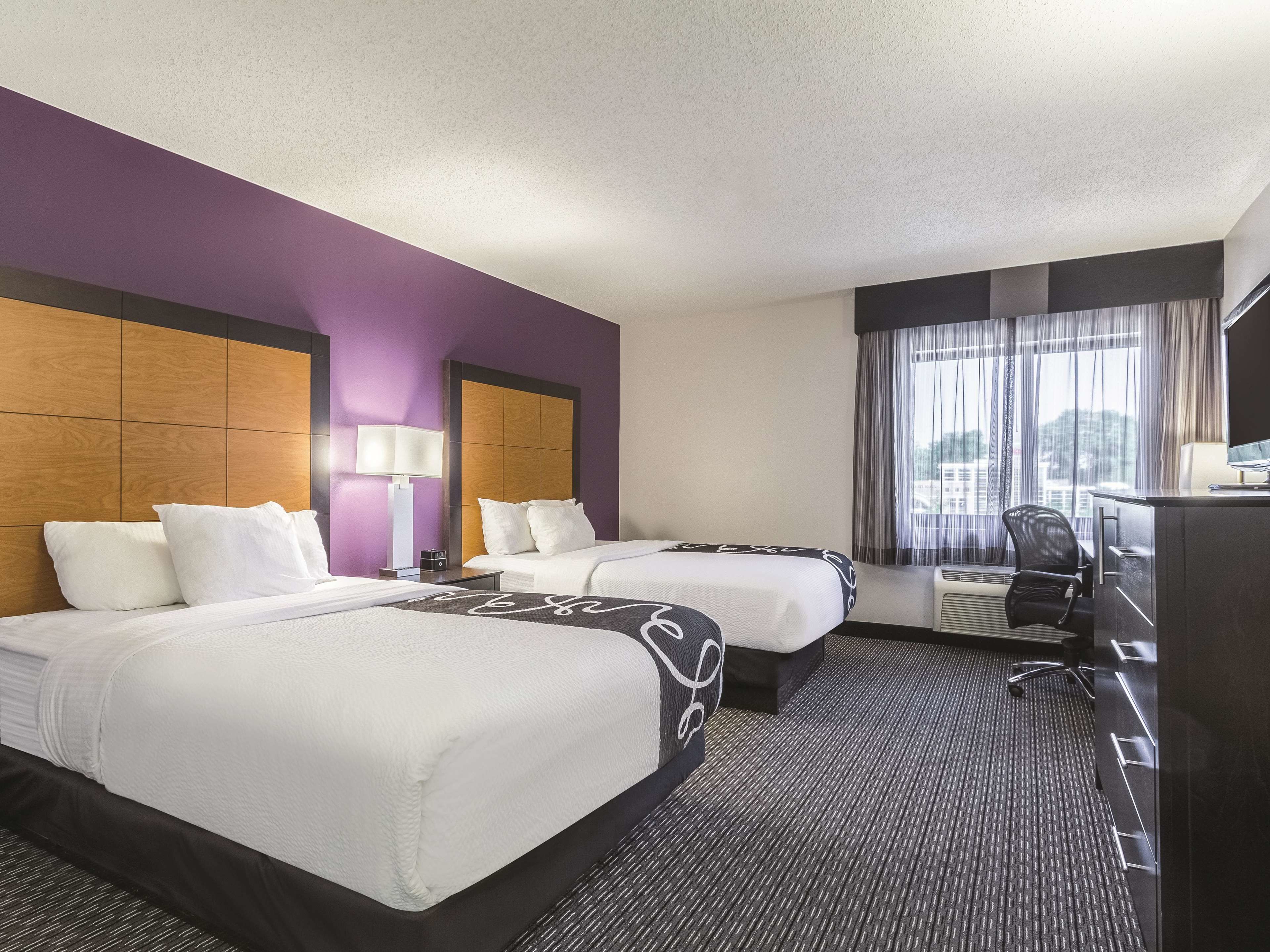 A hotel room with two beds, a desk, a chair, a lamp, and a view of the city through the window.