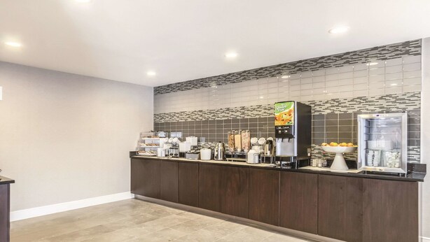Zona de recepción de un hotel con un mostrador de madera oscura, una máquina de café, un frutero y un refrigerador.