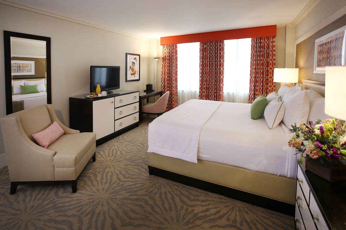 A hotel room with a bed, a desk with a television, a chair, and a desk lamp.