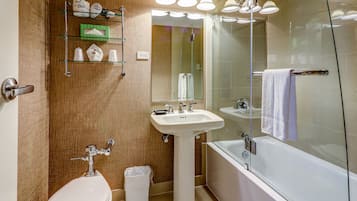 Un baño con lavamanos, inodoro y bañera. Hay repisas sobre el lavamanos y un bote de basura al lado del inodoro.