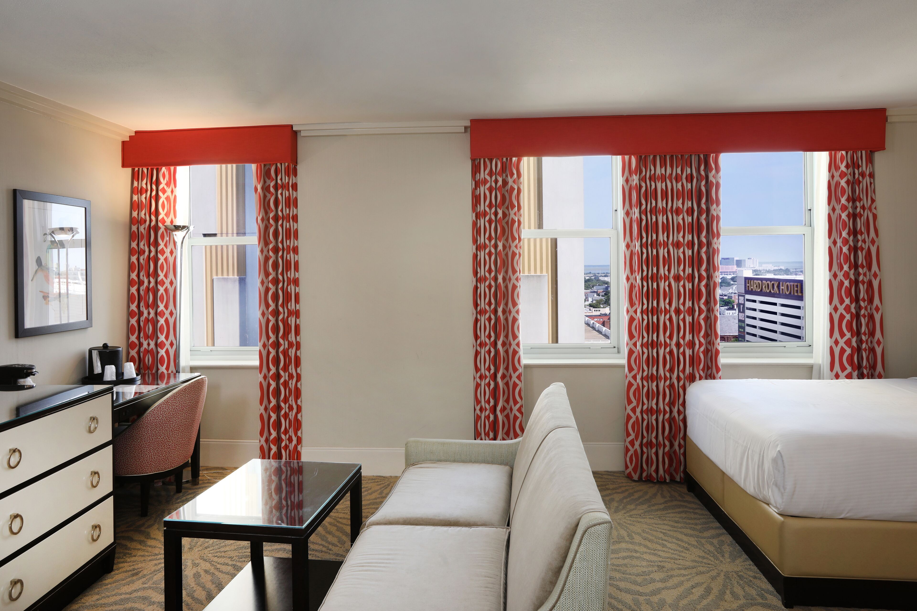 Ein Hotelzimmer mit einem Bett, einem Sofa, einem Schreibtisch und einem Stuhl. Es gibt Fenster mit Vorhängen und Blick auf die Stadt.