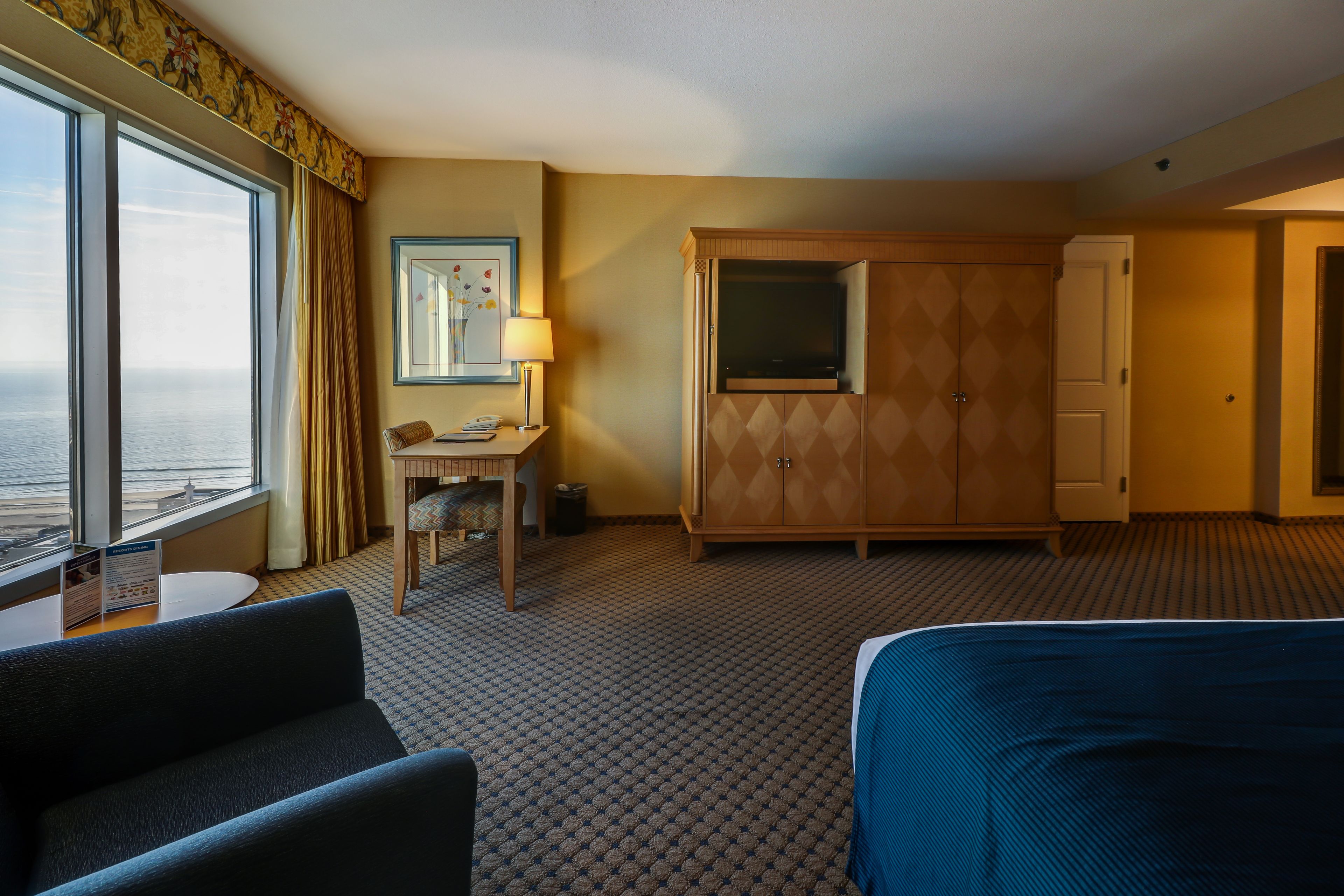 A hotel room with a large window, a desk with a lamp, a wardrobe, and a bed.