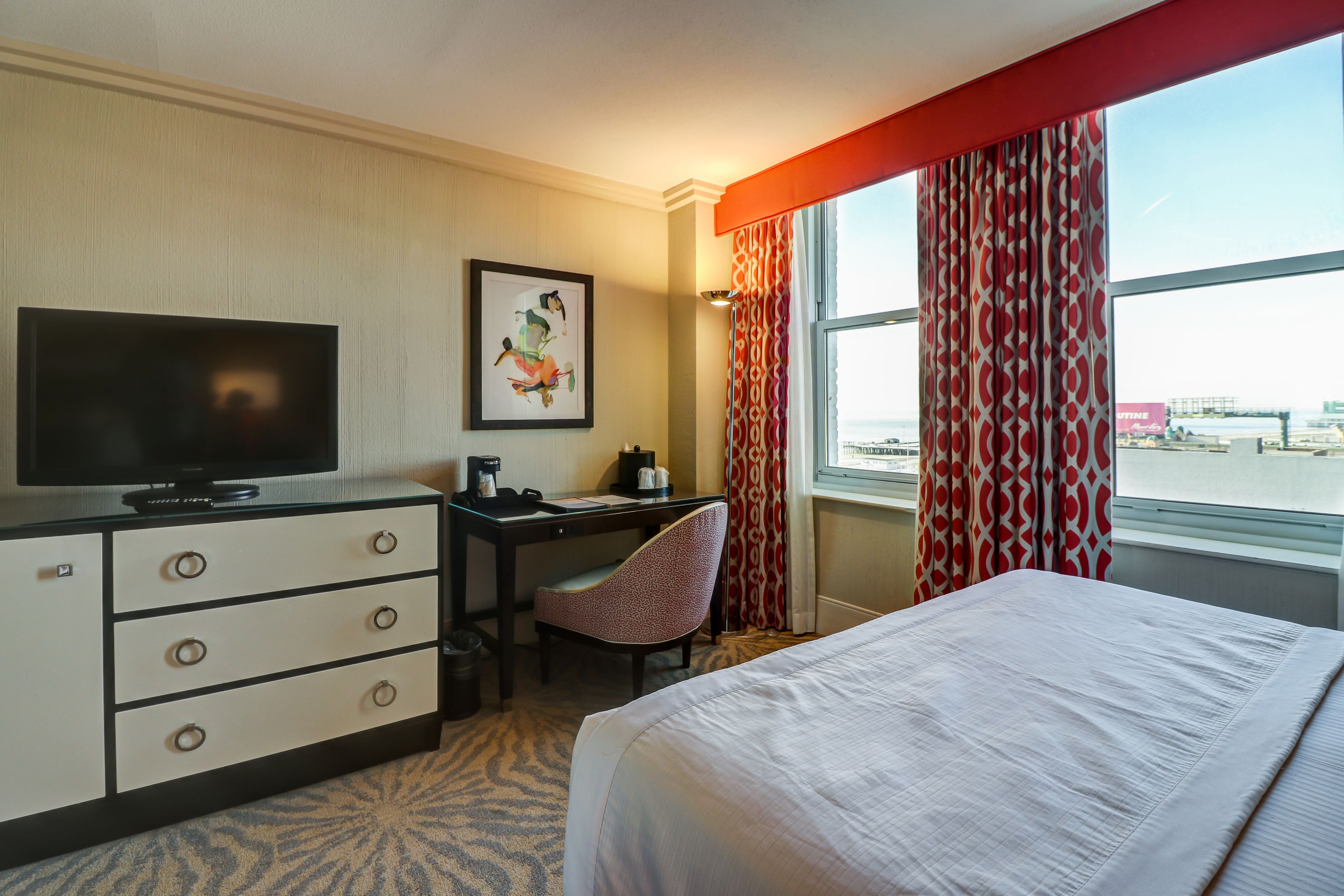 A hotel room with a bed, a desk with a chair, a television, and a window with curtains.