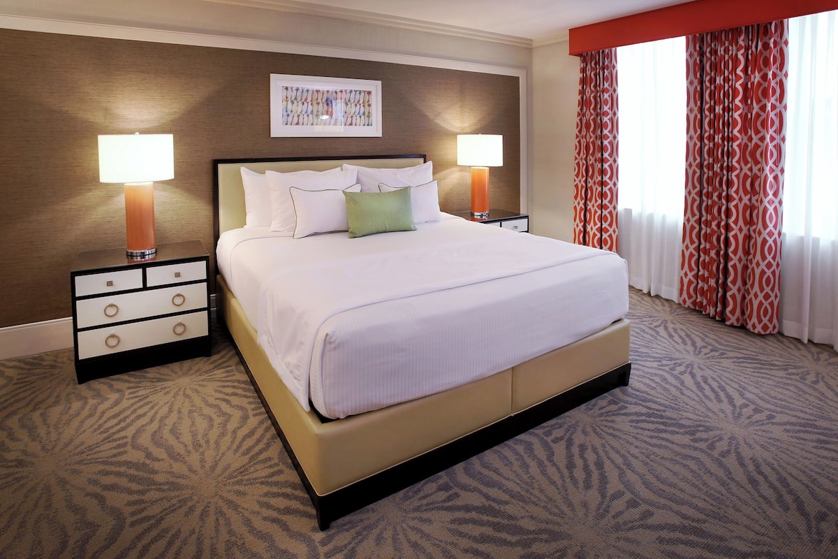 A hotel room with a large bed, two bedside tables with lamps, a carpeted floor, and a window with curtains.