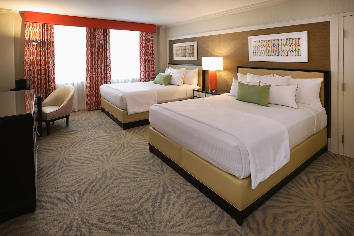 A hotel room with two beds, a desk, a chair, and a large window with curtains.