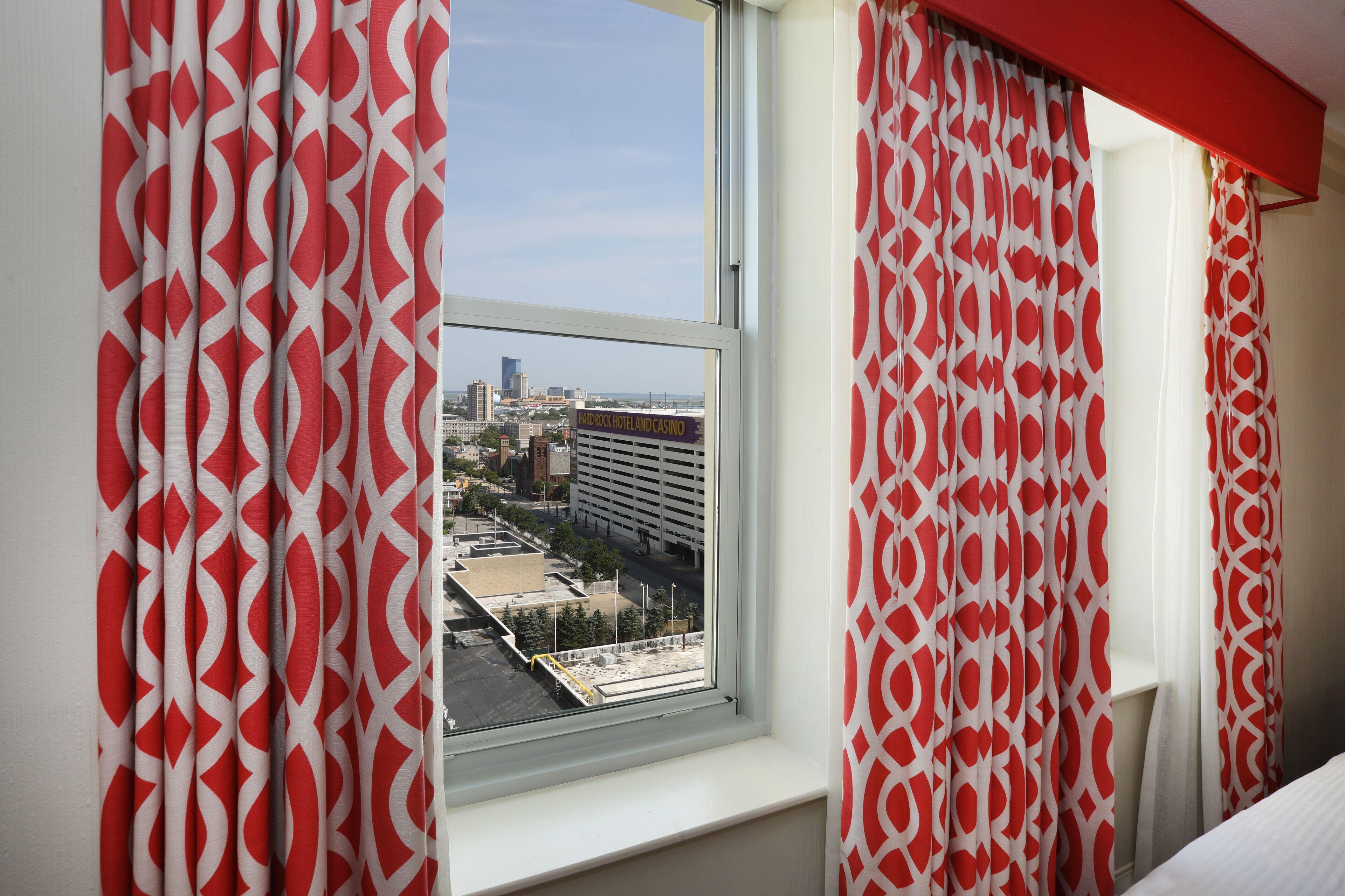 A hotel room with a large window, red and white patterned curtains, and a view of the cityscape.