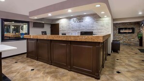 A reception area with a marble counter, dark wooden cabinets, a stone wall with a fireplace, and a mounted television. - Baymont by Wyndham Lancaster (Lancaster)