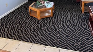 A living room with a patterned carpet, a glass coffee table, and several potted plants on a windowsill. - Budgetel Inn & Suites (Columbus)