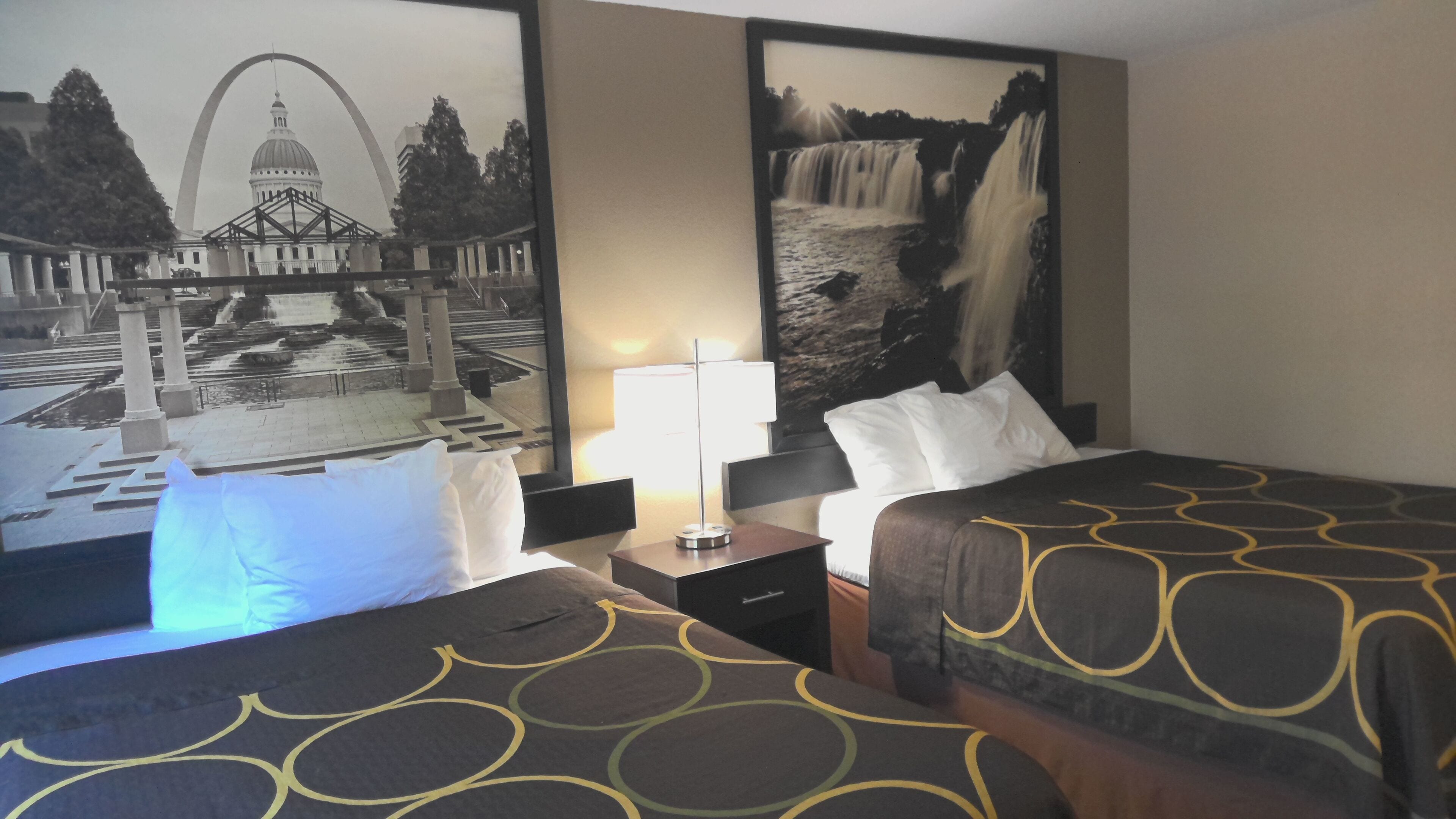 A hotel room with two beds, a nightstand, a lamp, and two large wall murals depicting a cityscape and a waterfall.