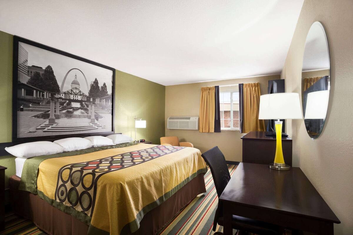 Een hotelkamer met een bed, een bureau, een stoel, een lamp, een raam met gordijnen, en een groot fotokader aan de muur.