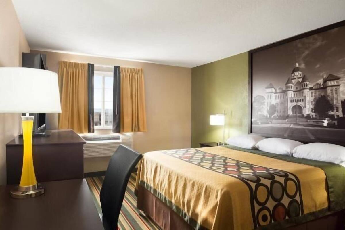 Een hotelkamer met een bed, een bureau met lamp, een stoel, een raam met gordijnen en een aan de muur hangend schilderij.