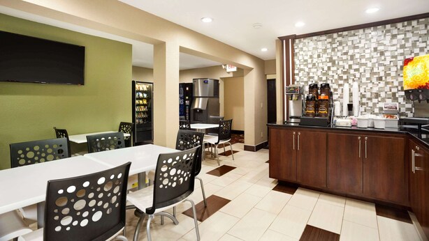 A hotel dining area with tables and chairs, a kitchen area with a coffee station, and a flat-screen TV mounted on the wall. - Super 8 by Wyndham Joplin (Joplin)