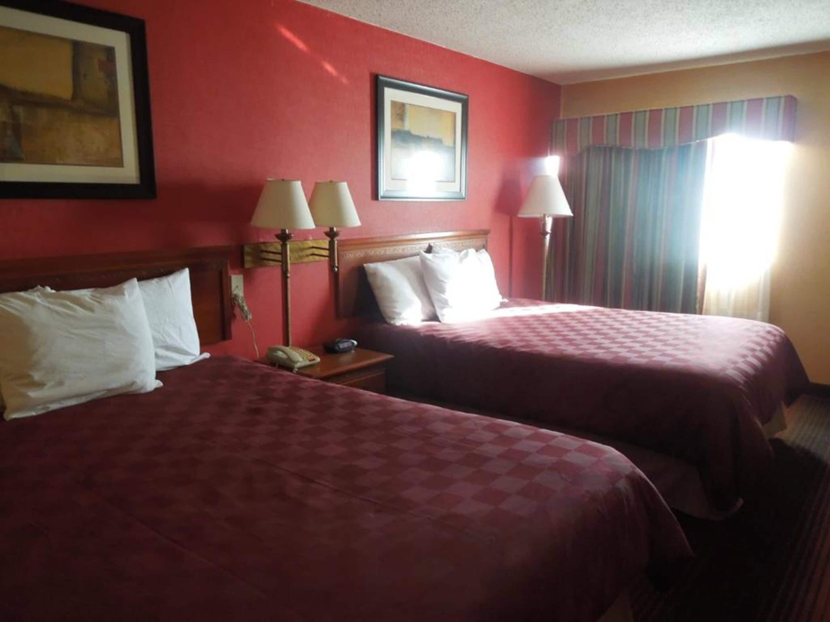 A hotel room with two beds, a nightstand with a lamp, a framed picture on the wall, and a window with curtains.