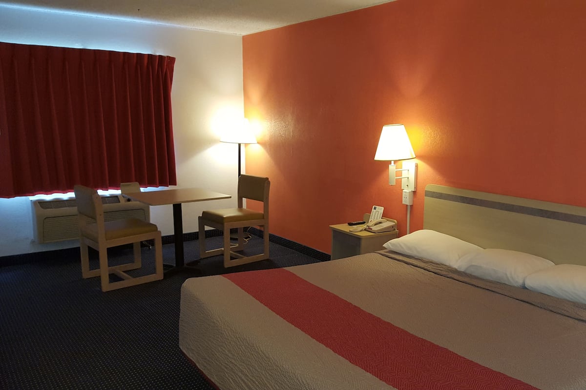 Quarto de hotel com uma cama, uma escrivaninha, duas cadeiras, um telefone e duas luminárias de parede.