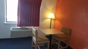 Ein kleines Hotelzimmer mit einem Tisch und zwei Stühlen, einem Fenster mit roten Vorhängen, einer Wandlampe und einer Heizung.