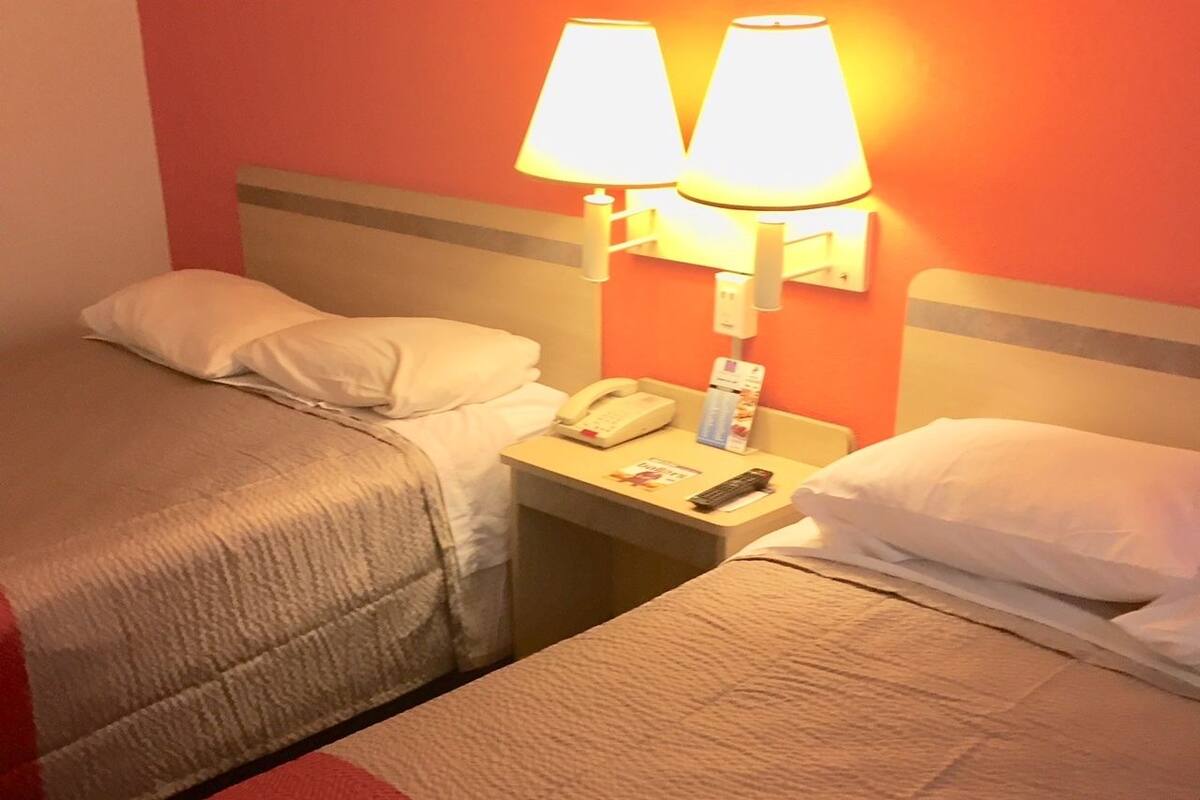 Quarto de hotel com duas camas, mesa de cabeceira com telefone e abajur, e luminária na parede.