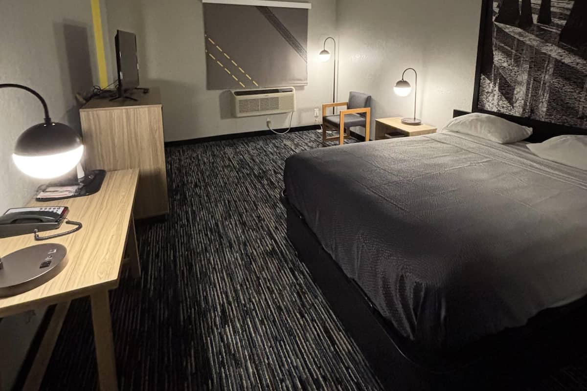A hotel room with a bed, desk, chair, lamp, air conditioning unit, and a wall-mounted TV.