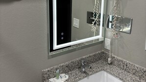 A bathroom with a granite countertop, a white sink, a mirror with a light, and a hairdryer mounted on the wall. - Super 8 Springfield North I-44 (Springfield)