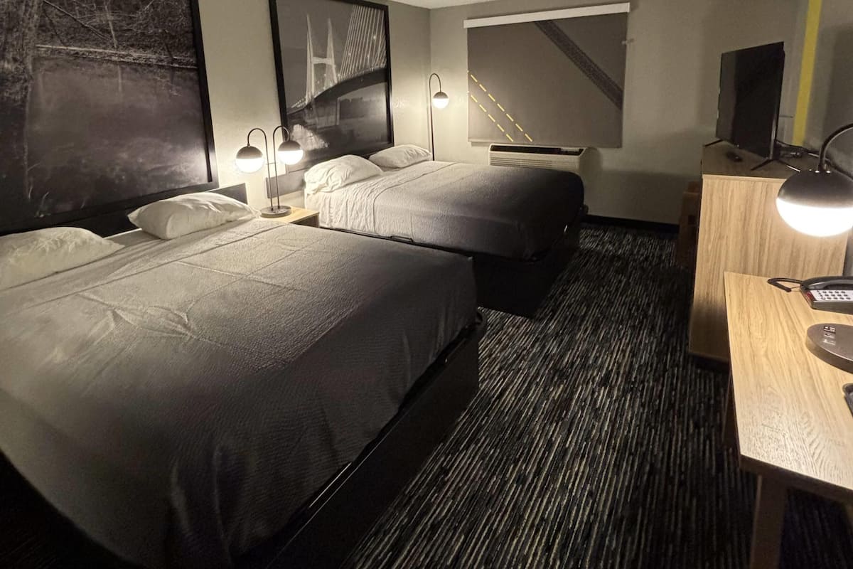 A hotel room with two beds, a desk, a chair, a TV, and a large window with a view of a bridge.