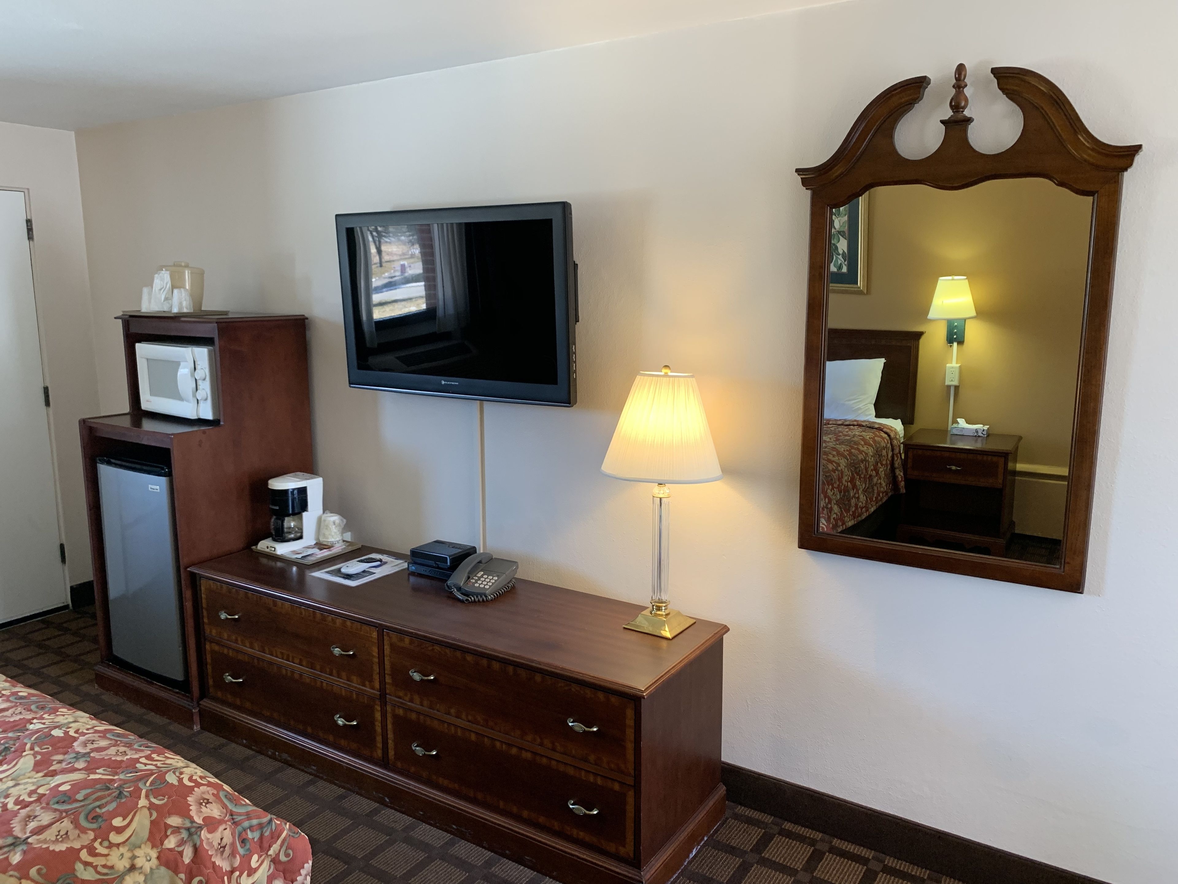 A hotel room with a television, a bedside lamp, a mirror, a minibar, and a bed.