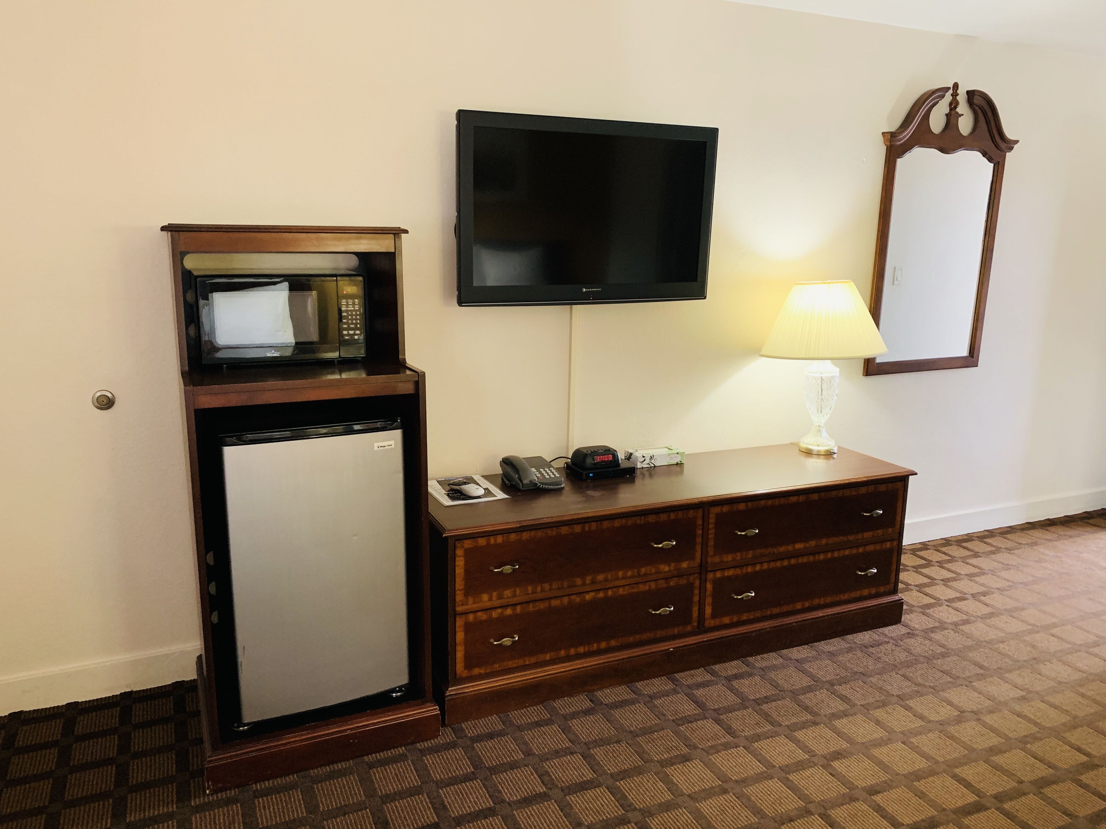 A hotel room with a minibar, a flat-screen TV, a desk with a lamp, and a mirror.