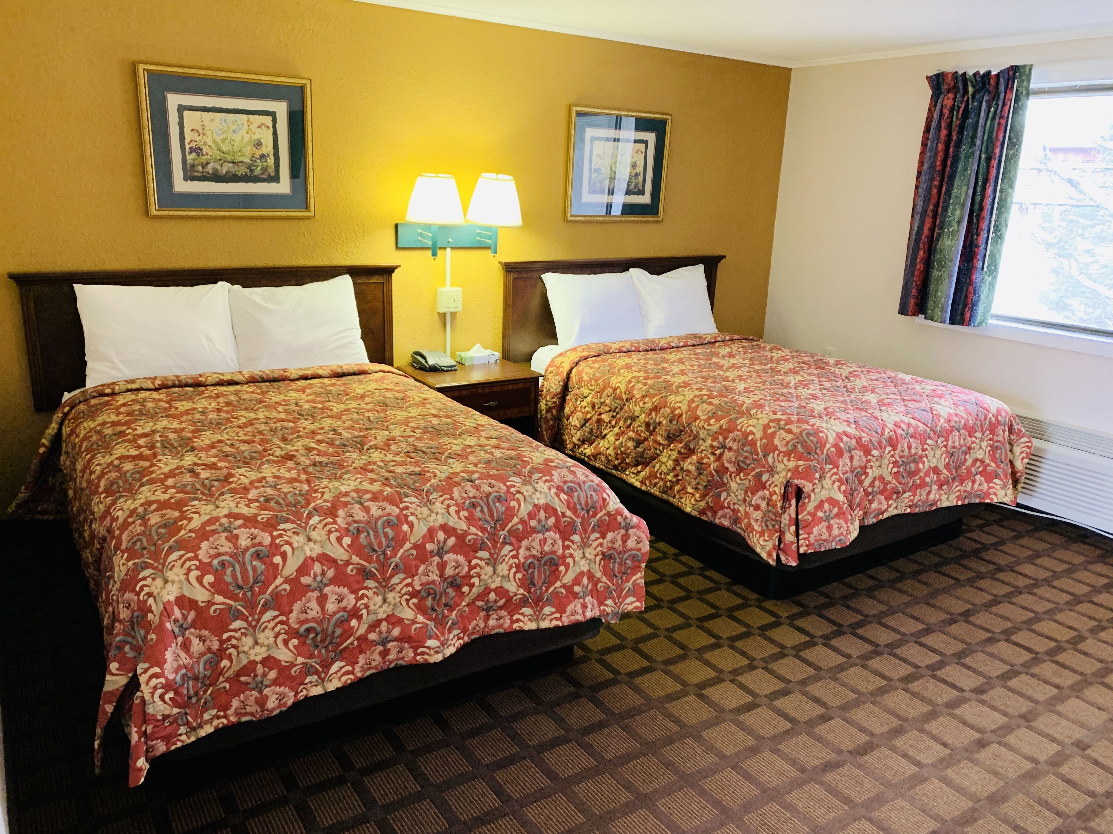 A hotel room with two beds, a nightstand with a lamp, framed pictures on the wall, and a window with curtains.