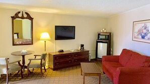 A hotel room with a bed, a TV, a desk with a lamp, a microwave, a refrigerator, a red sofa, and a painting on the wall.