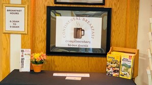 A reception area with a framed welcome sign, a no smoking sign, a breakfast hours sign, a continental breakfast sign, and travel guides on the table.