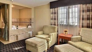A hotel room with a bathroom, two armchairs, a small table, and a large window with curtains.