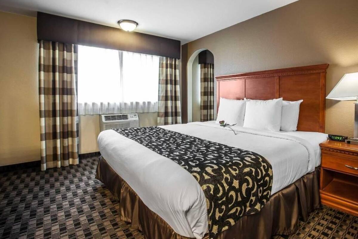 A hotel room with a large bed, a wooden headboard, a nightstand, a lamp, and a window with curtains.