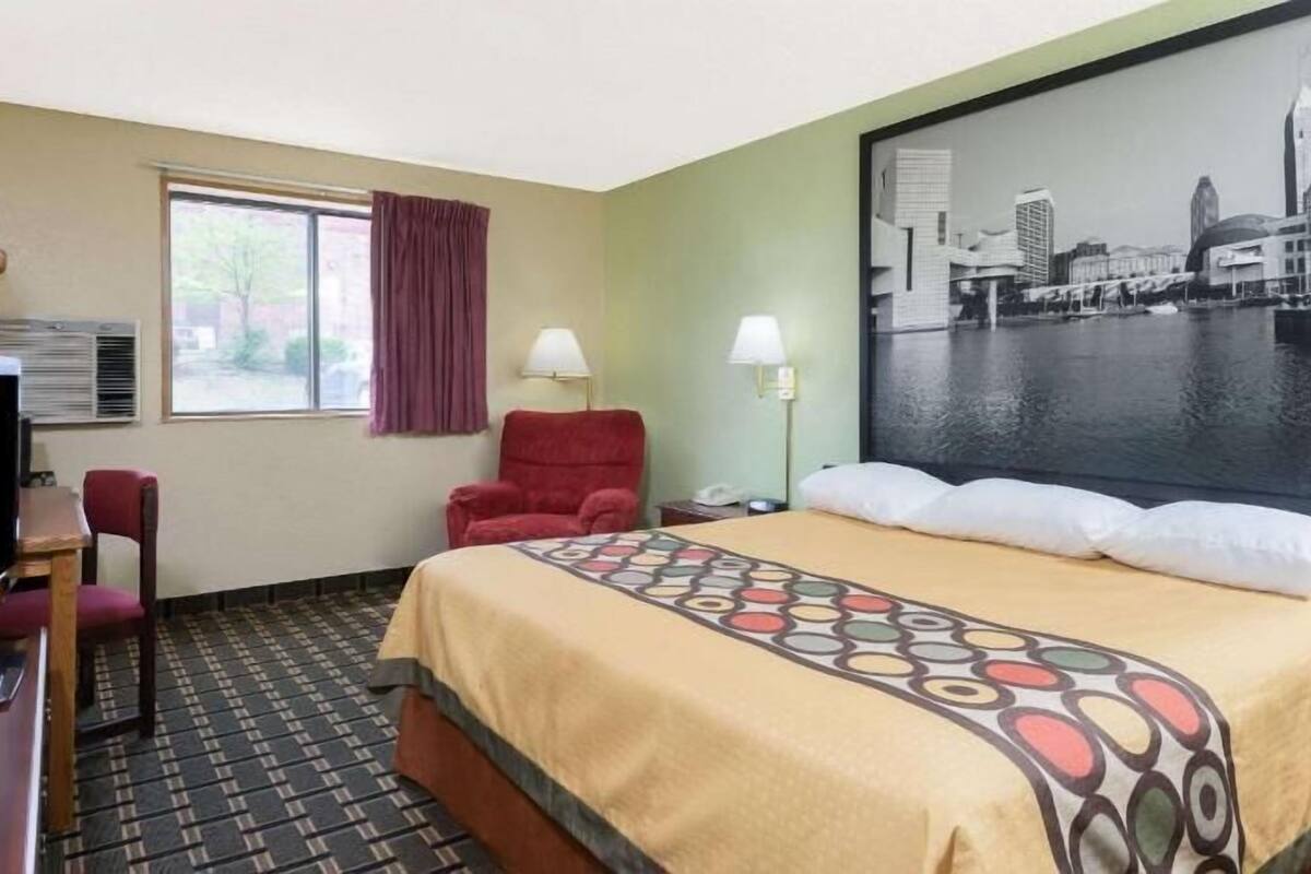 Een hotelkamer met een bed, een bureau met een televisie, een rode fauteuil, een raam met gordijnen en een stadsgezichtschilderij aan de muur.