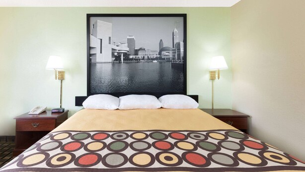 Una habitación de hotel con una cama, dos mesitas de noche, una lámpara de noche, una obra de arte con un paisaje urbano y dos lámparas.
