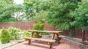 Una mesa de picnic de madera con bancos en un jardín ajardinado, con un cerco de madera y diversos arbustos.