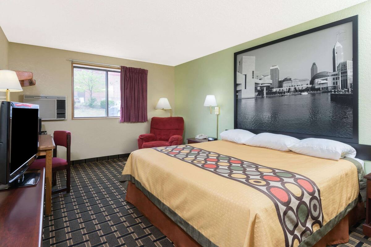 Een hotelkamer met een bed, een bureau met een televisie, een stoel, een raam met gordijnen en een stadslandschap schilderij aan de muur.