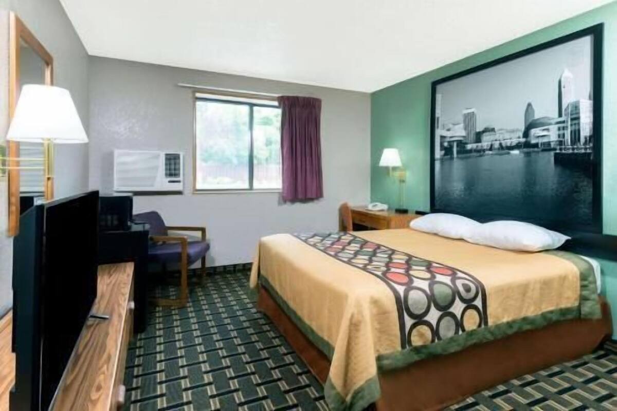 Een hotelkamer met een bed, een bureau, een stoel, een televisie, een raam met gordijnen en een stadslandschap schilderij aan de muur.