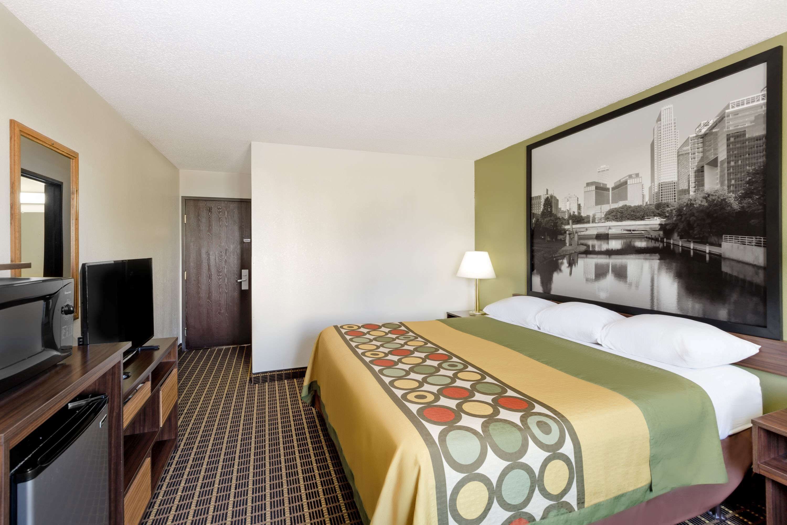 A hotel room with a bed, a TV, a desk, and a large window with a cityscape view.