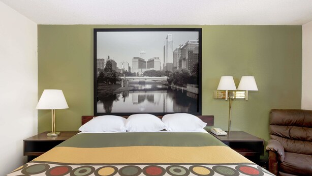 A hotel room with a bed, two bedside lamps, a brown leather chair, and a cityscape painting on the wall. - Super 8 by Wyndham Norfolk (Norfolk)
