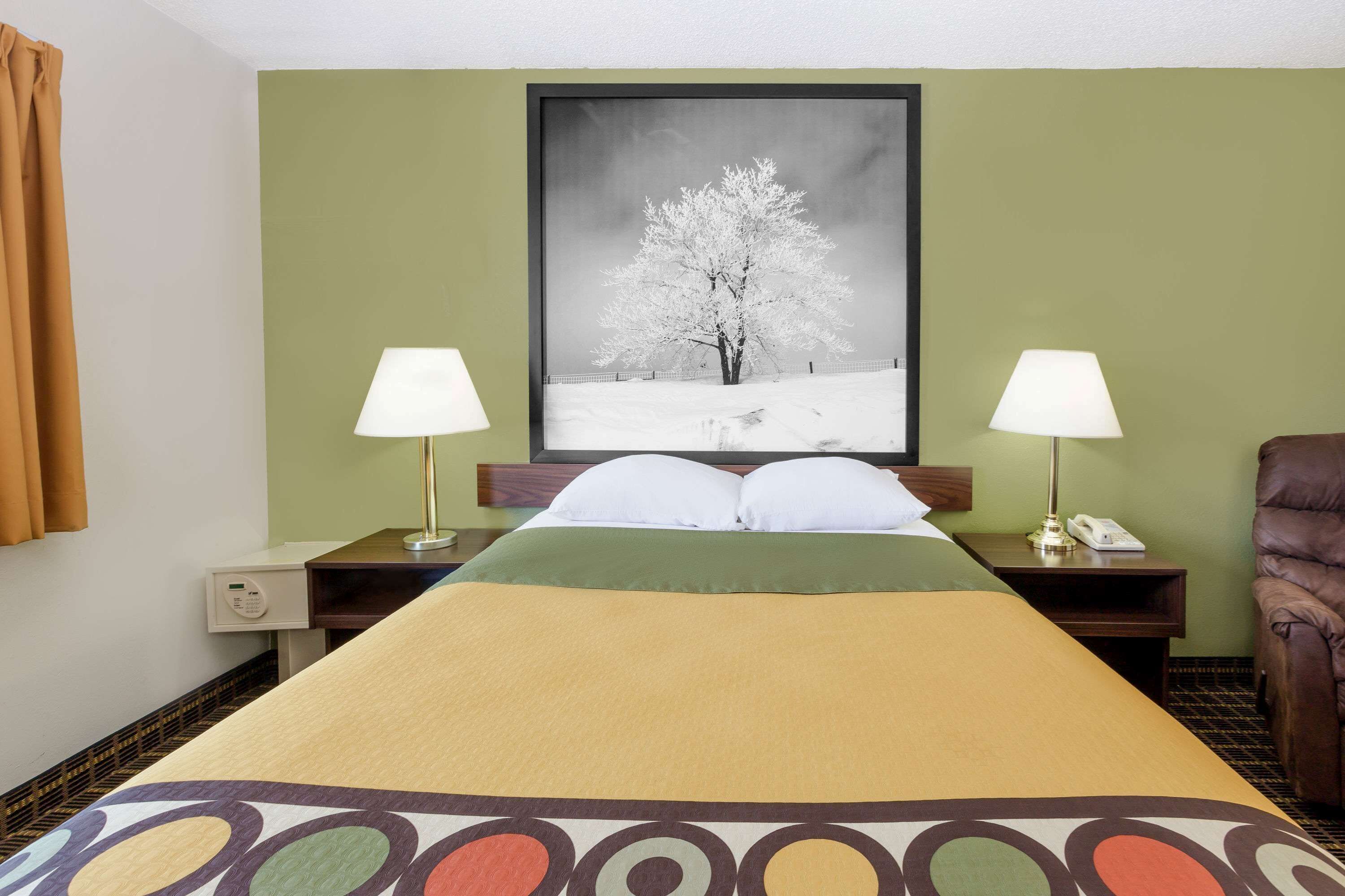 Una habitación de hotel con una cama, dos mesitas de noche con lámparas, una silla, un cuadro en la pared y una ventana con cortinas.