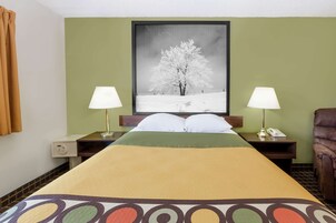Una habitación de hotel con una cama, dos mesitas de noche con lámparas, una silla, un cuadro en la pared y una ventana con cortinas.