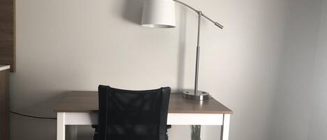 Un escritorio pequeño y blanco con una silla de oficina negra y una lámpara de escritorio blanca.