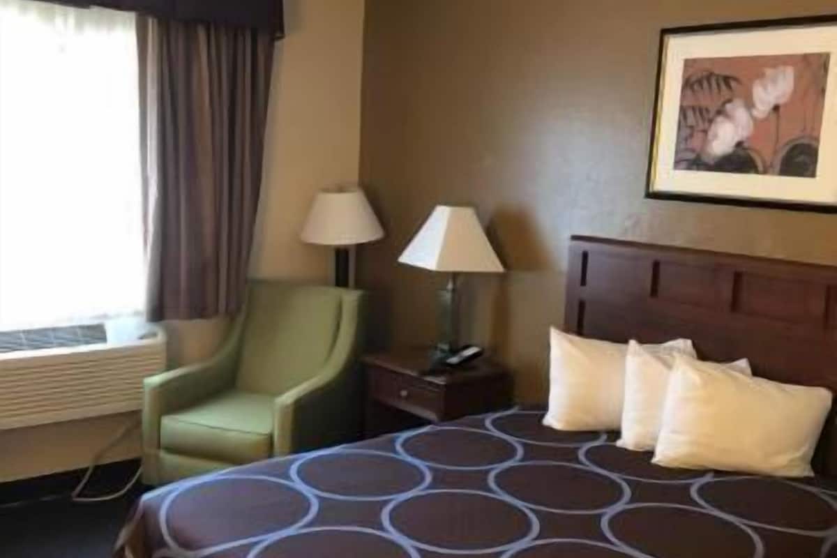 Een hotelkamer met een bed, een stoel, een nachtkastje, een lamp, een foto aan de muur en een raam met gordijnen.
