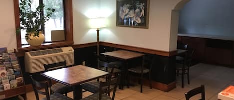 Interior de restaurante con mesas y sillas, una ventana con una planta, un cuadro colgado en la pared y piso de baldosas.