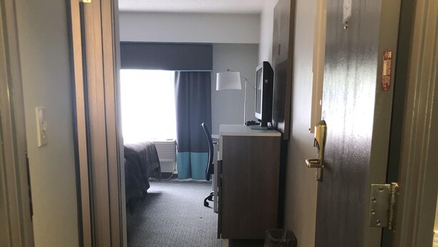 Pasillo de hotel con piso alfombrado, un escritorio con lámpara, televisión y una cama al fondo.