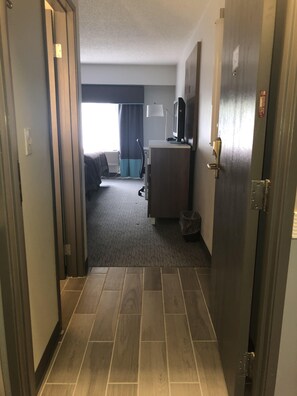 Pasillo de hotel con piso alfombrado, un escritorio con lámpara, televisión y una cama al fondo.