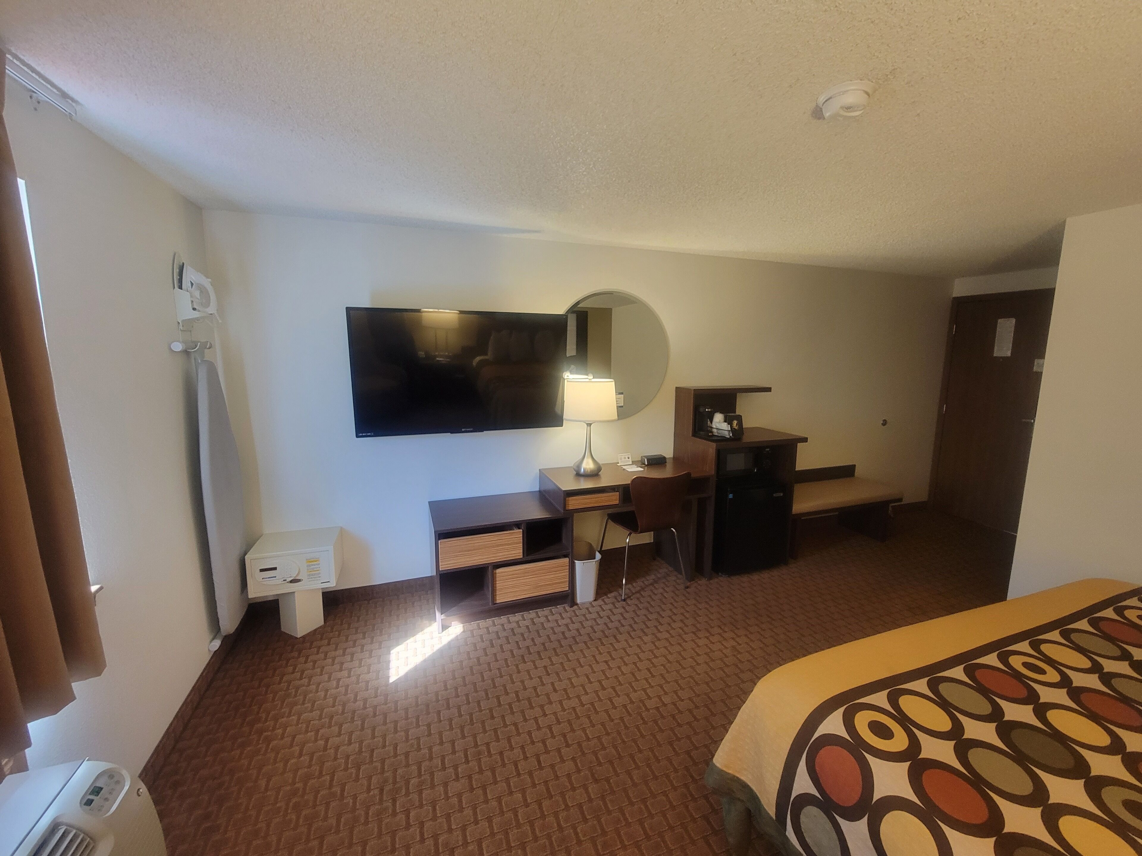A hotel room with a flat-screen TV, a desk with a lamp, a small refrigerator, a bench, and a patterned bedspread.