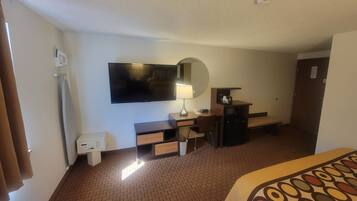 A hotel room with a flat-screen TV, a desk with a lamp, a small refrigerator, a bench, and a patterned bedspread.